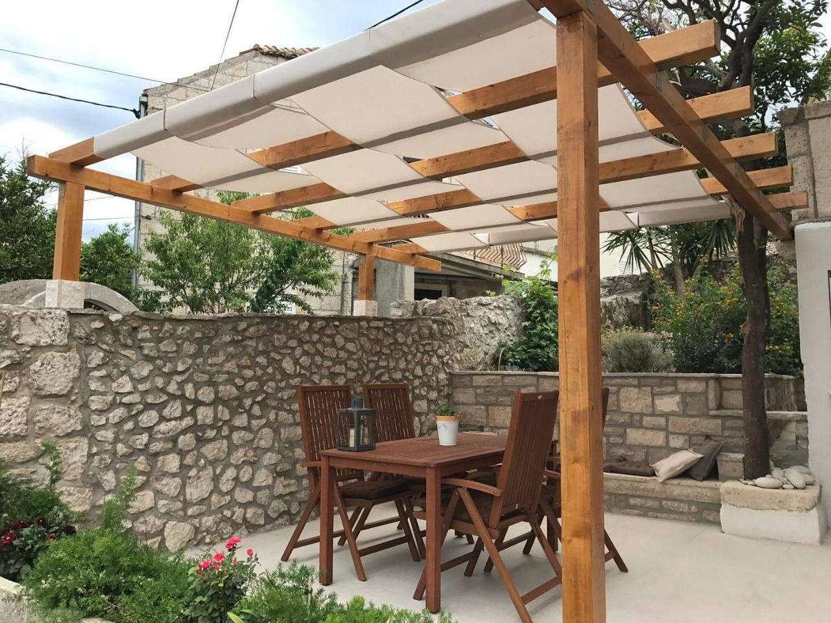 Wooden pergola with cream fabric canopy over outdoor dining area, stone wall garden